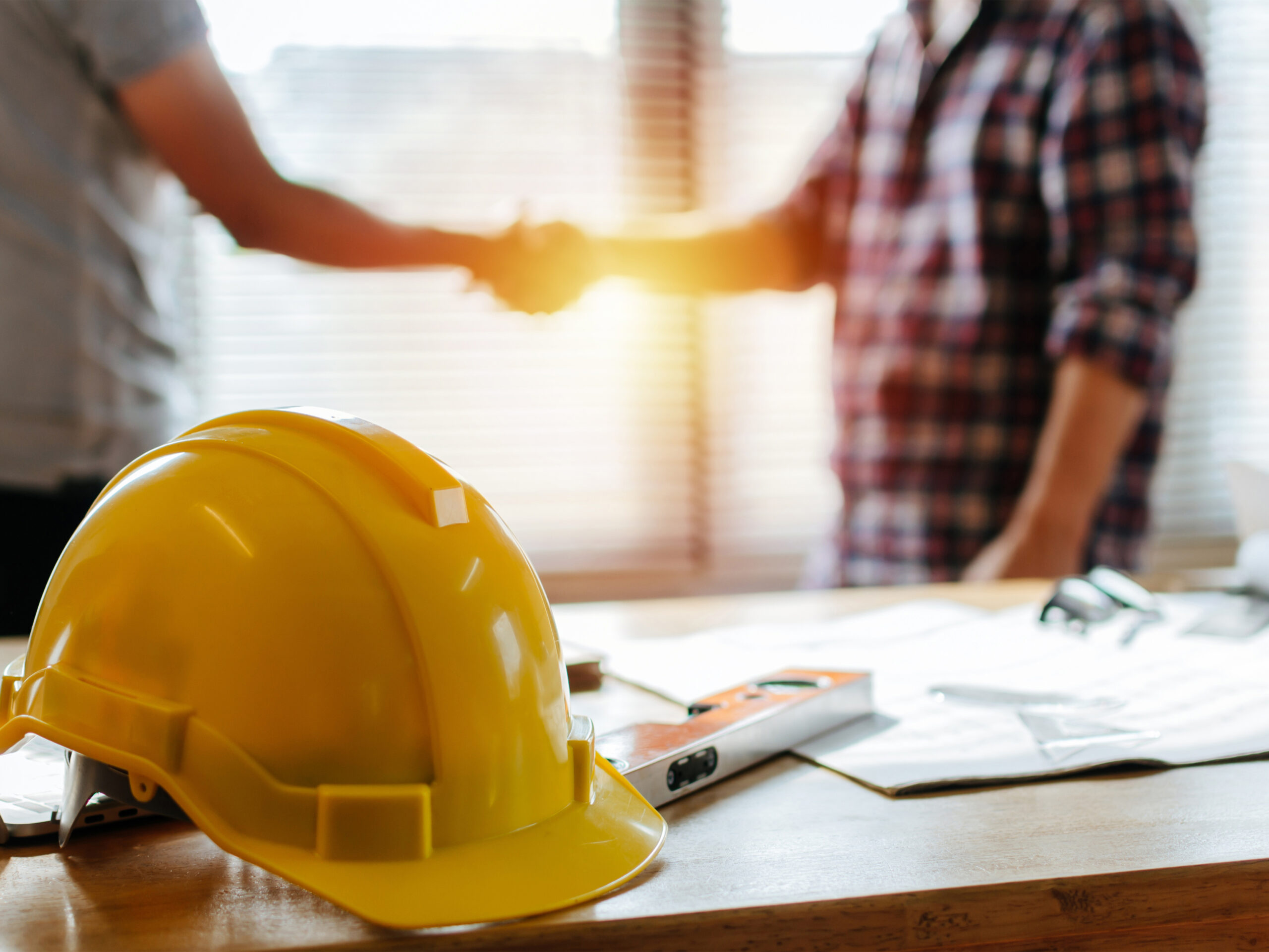 yellow safety helmet on workplace desk with construction worker team hands shaking greeting start up plan new project contract in office center at construction site, partnership and contractor concept
